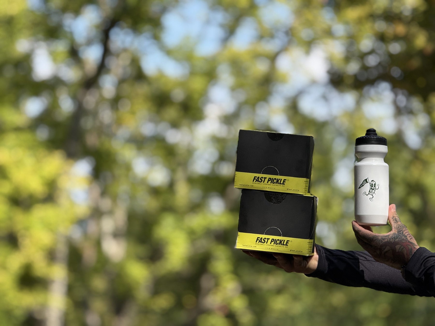 Two Fast Pickle 12-count boxes and a white Fast Pickle water bottle held up against green trees
